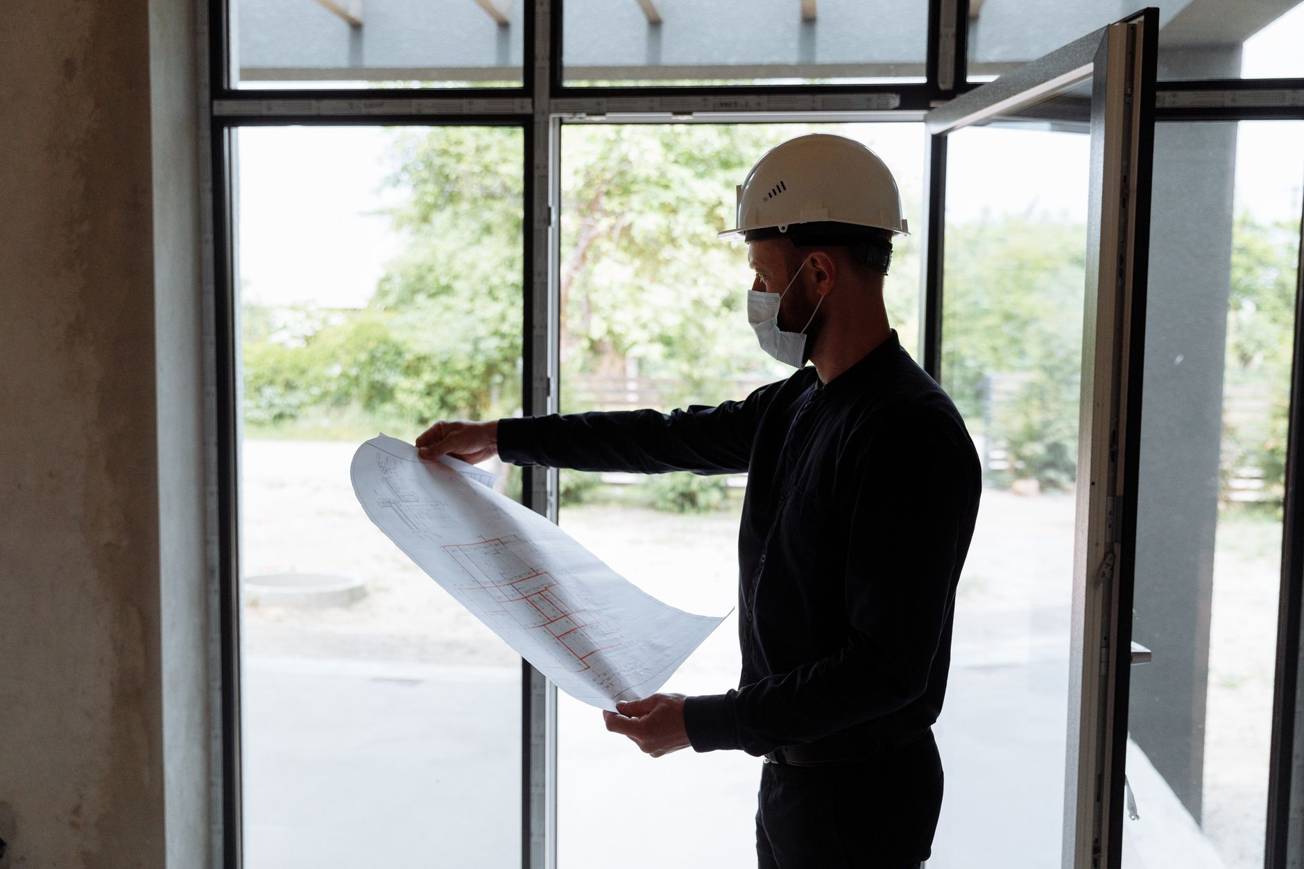Architect reviewing floor plans indoors, wearing safety gear, focused on construction planning.