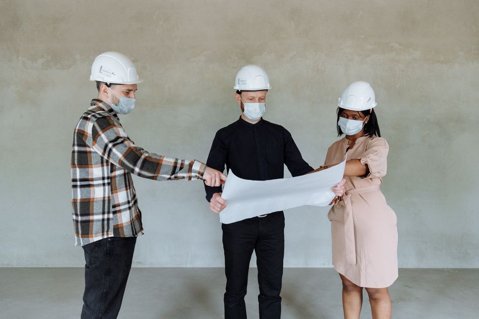 Tres arquitectos llevan máscaras y cascos mientras revisan planos de construcción en interiores.