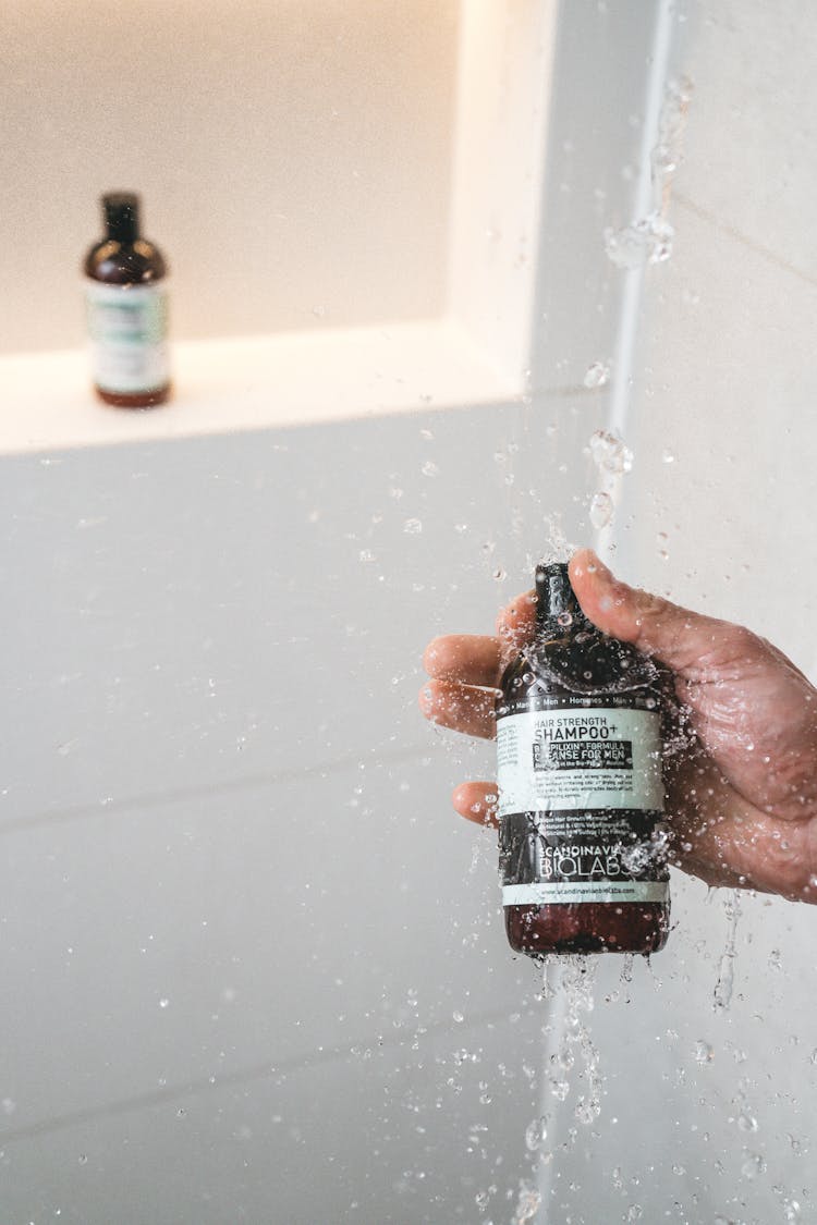 Hand Holding Bottle Of Shampoo Under Shower Water