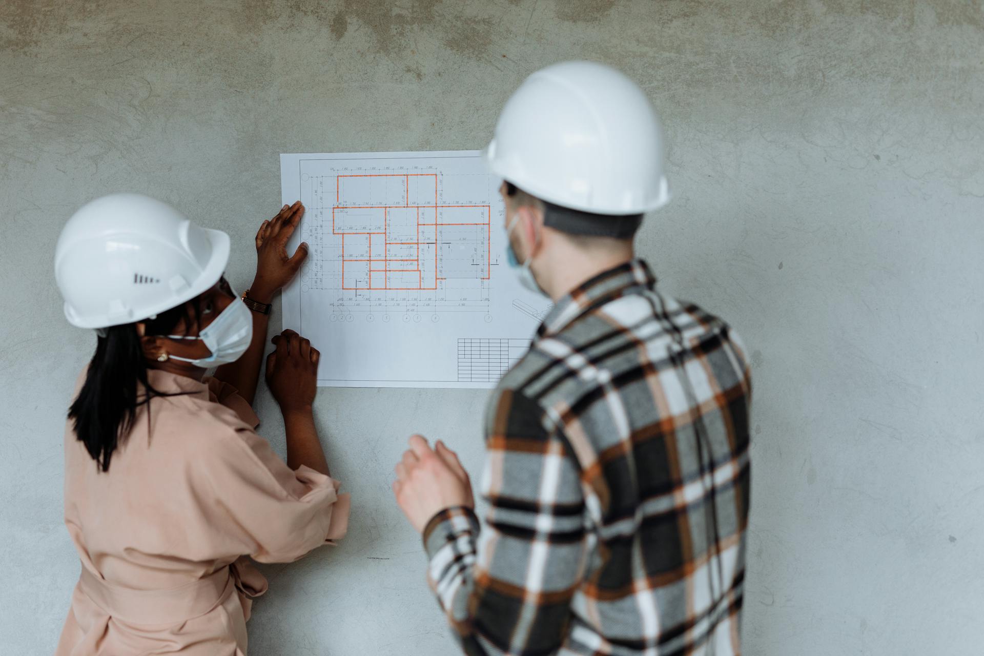 Two architects in hard hats and masks review blueprints during a building project.