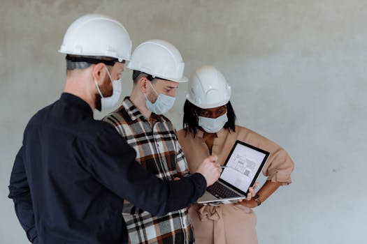 Group of diverse architects with safety gear examining building plans on a laptop.
