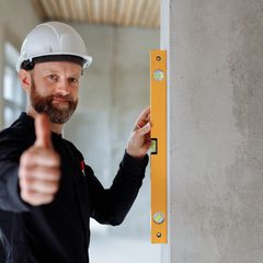 Smiling builder in a white hard hat giving a thumbs up while holding a yellow spirit level upright against a wall