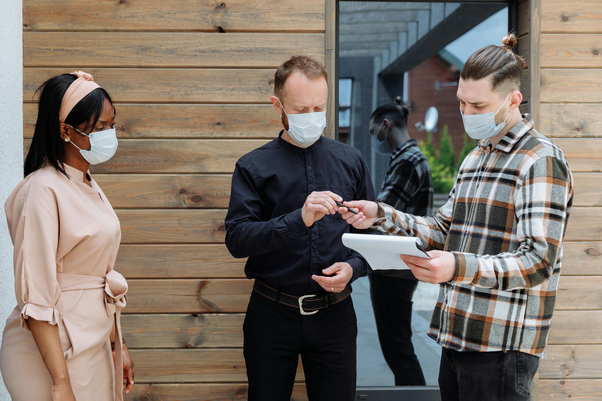 Three adults signing a real estate contract outdoors, all wearing face masks for safety.