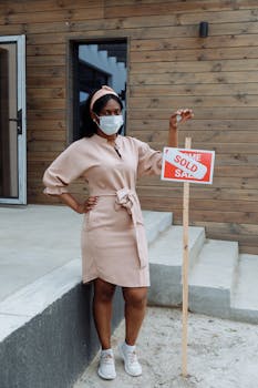 Woman wearing a face mask holds a 'Sold' sign outside a modern wooden house.