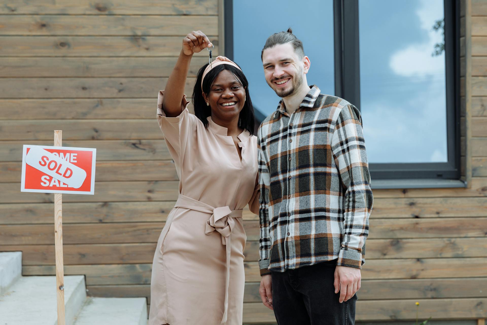 Young couple celebrates buying their first house with keys in hand and a sold sign.