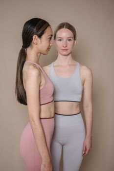Two women in sports bras and tights standing against a beige background.