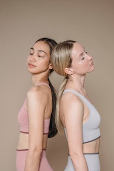 Two women in activewear with closed eyes posing back to back against a beige studio background.