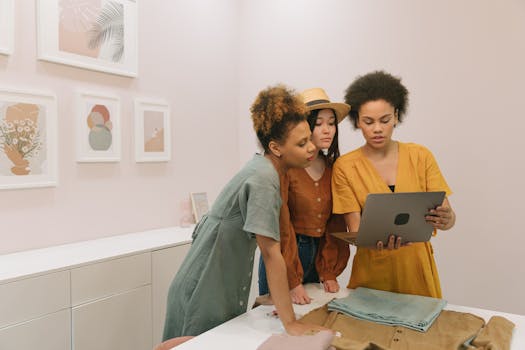 Three diverse women stand indoors, collaborating on a laptop surrounded by modern art.
