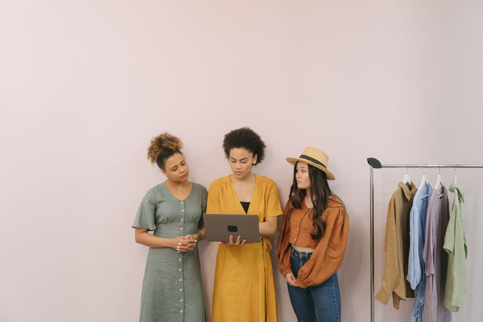 Três mulheres discutem estratégia de negócios em uma boutique de moda com roupas em exibição.