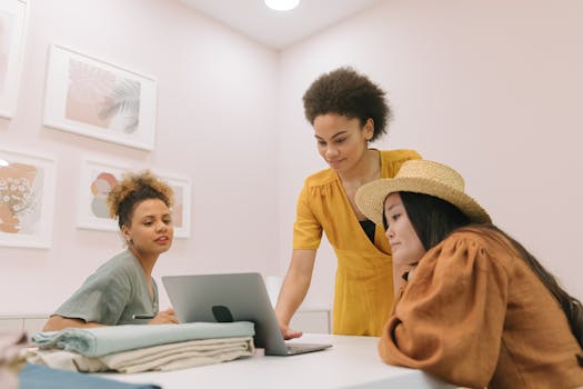 Three diverse women collaborating in a modern office setting with a laptop, showcasing teamwork.