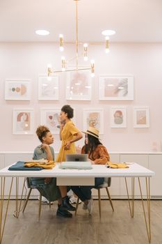 Three women discussing fashion design at a table with fabrics, laptop, and art decor indoors.