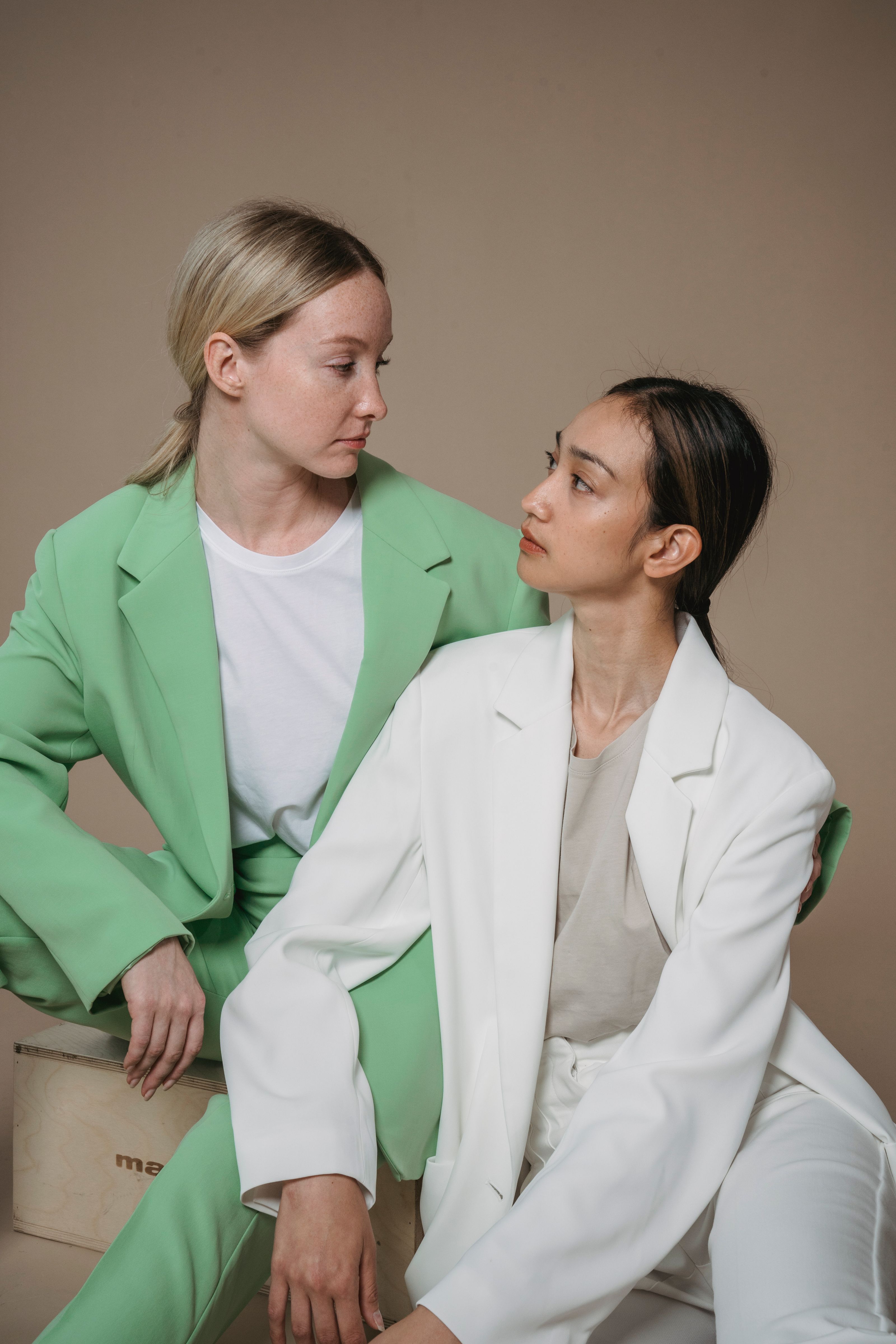 Duas mulheres em blazers pastel compartilham um momento em um retrato minimalista de estúdio.