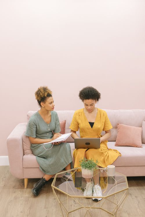 Free Two women sitting on a couch, working together with a laptop and notepad in a stylish indoor setting. Stock Photo
