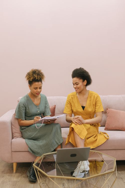 Free Two women collaborate on a project at home, sitting on a sofa with a laptop and documents. Stock Photo