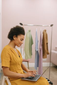 Woman in yellow dress using laptop in room with clothing rack. Ideal for remote work and fashion concepts.
