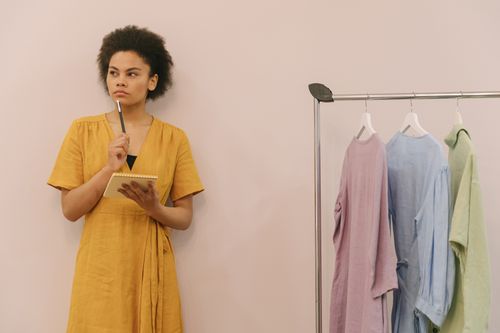 Free A woman in a yellow dress ponders creatively in a clothing studio with racks. Stock Photo