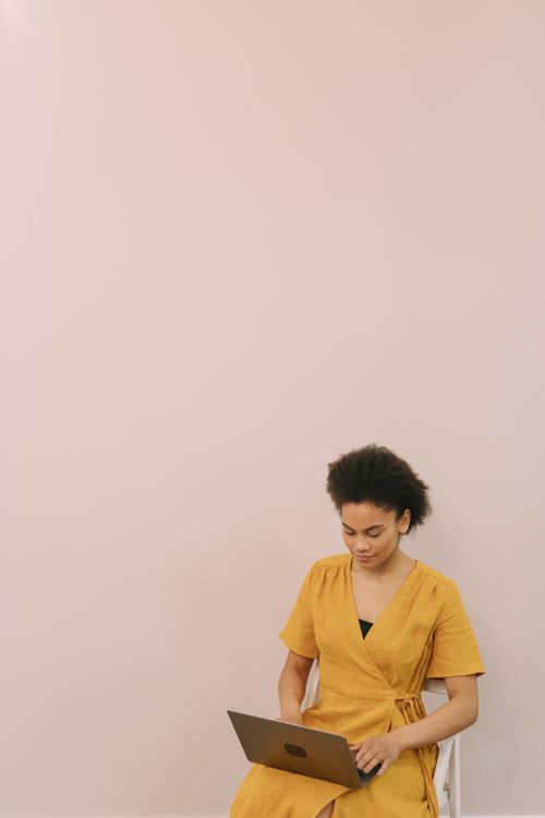 Free A woman in a yellow dress working on a laptop in a minimalist indoor setting. Stock Photo