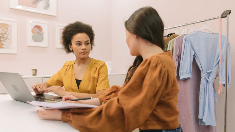 Two women discussing a fashion business strategy indoors with clothing rack.
