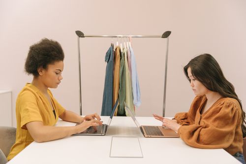 Free Two women focusing on their laptops in a modern office setting with a clothes rack in the background. Stock Photo