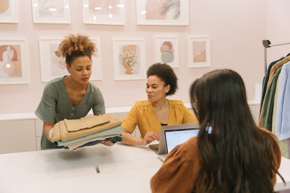 Três mulheres trabalhando juntas em uma boutique moderna, discutindo moda e estratégias de negócios.