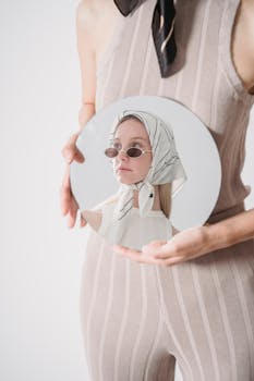 A stylish woman in sunglasses and scarf holding a round mirror reflecting her image.