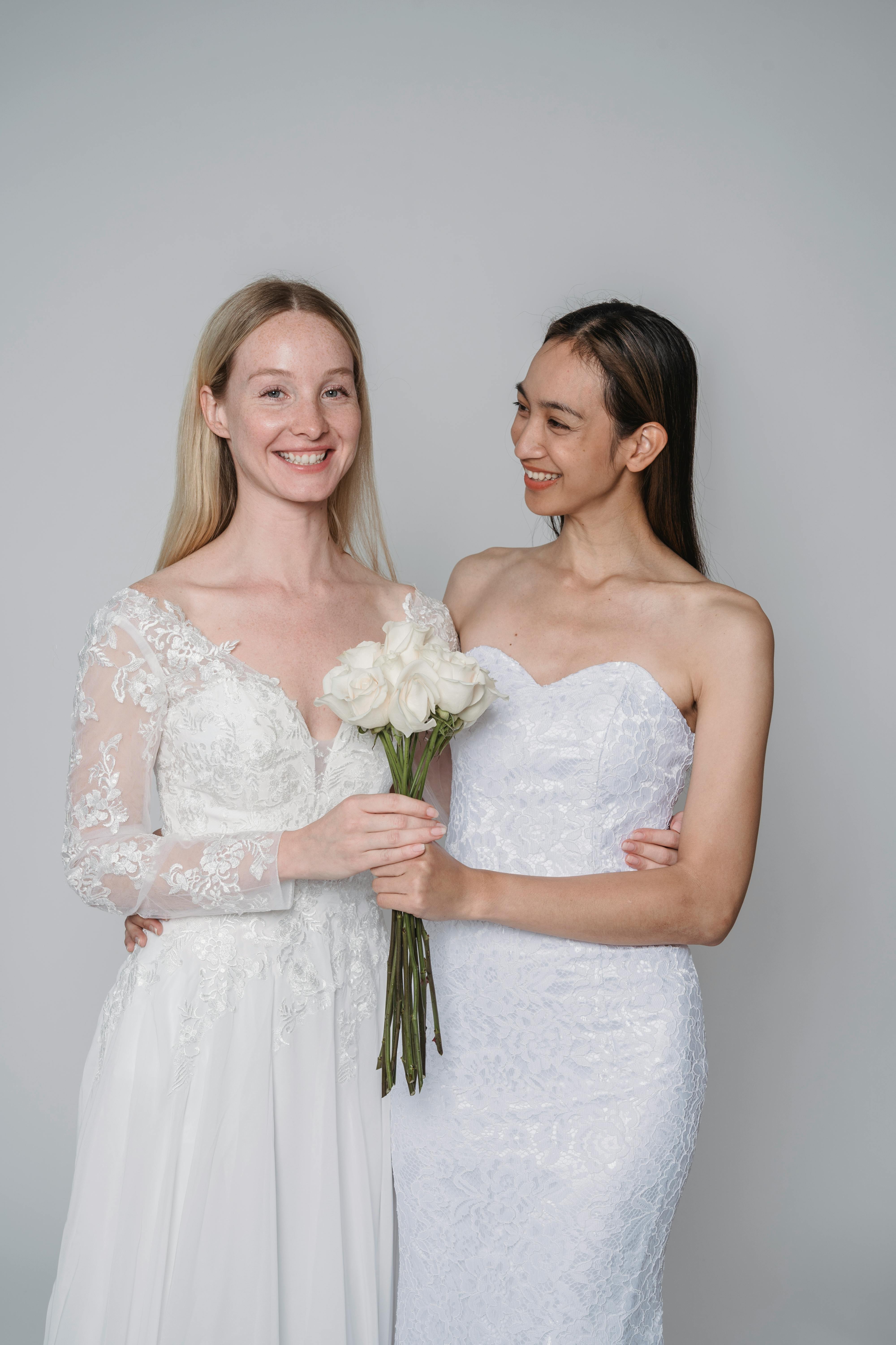 Two brides wearing elegant dresses with white bouquets, smiling in a studio setting.