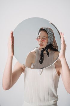 A woman in sunglasses and scarf holds a round mirror, creating an artistic reflection.