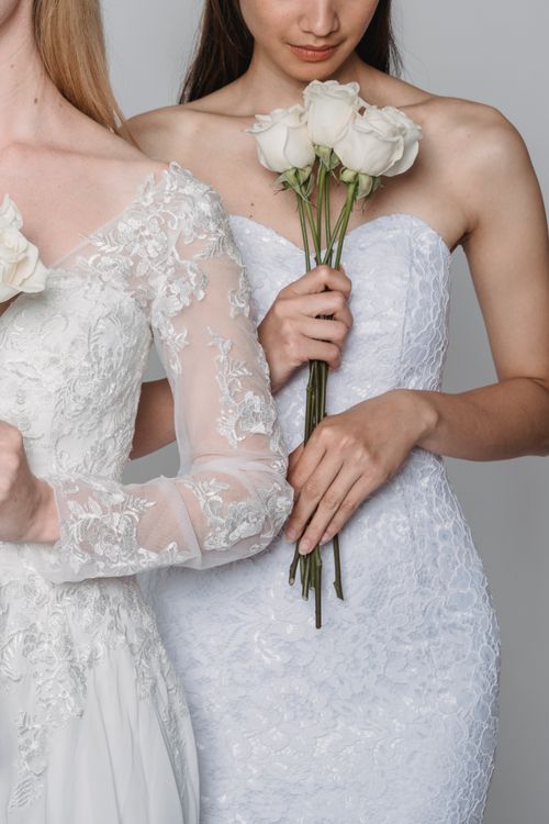 Free Two brides in intricately designed lace gowns pose gracefully, holding white roses. Stock Photo