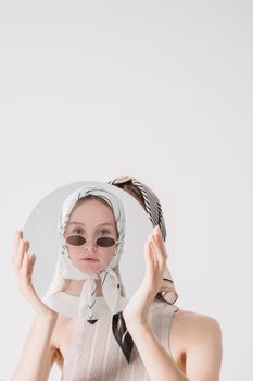 Elegant woman in headscarf and sunglasses holding a round mirror reflecting her own image.
