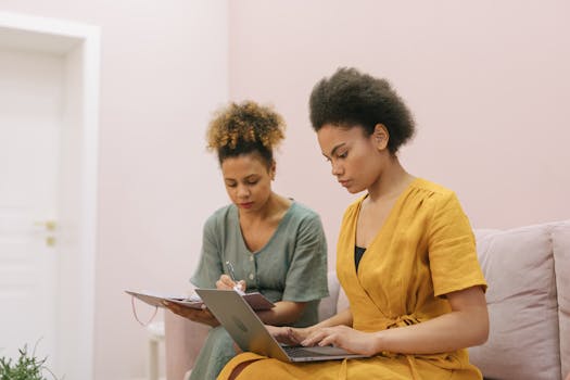 Two women collaborating in a modern office, using a laptop and taking notes.