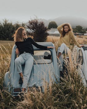 Stylish women in rustic setting pose on a vintage car, showcasing countryside fashion.