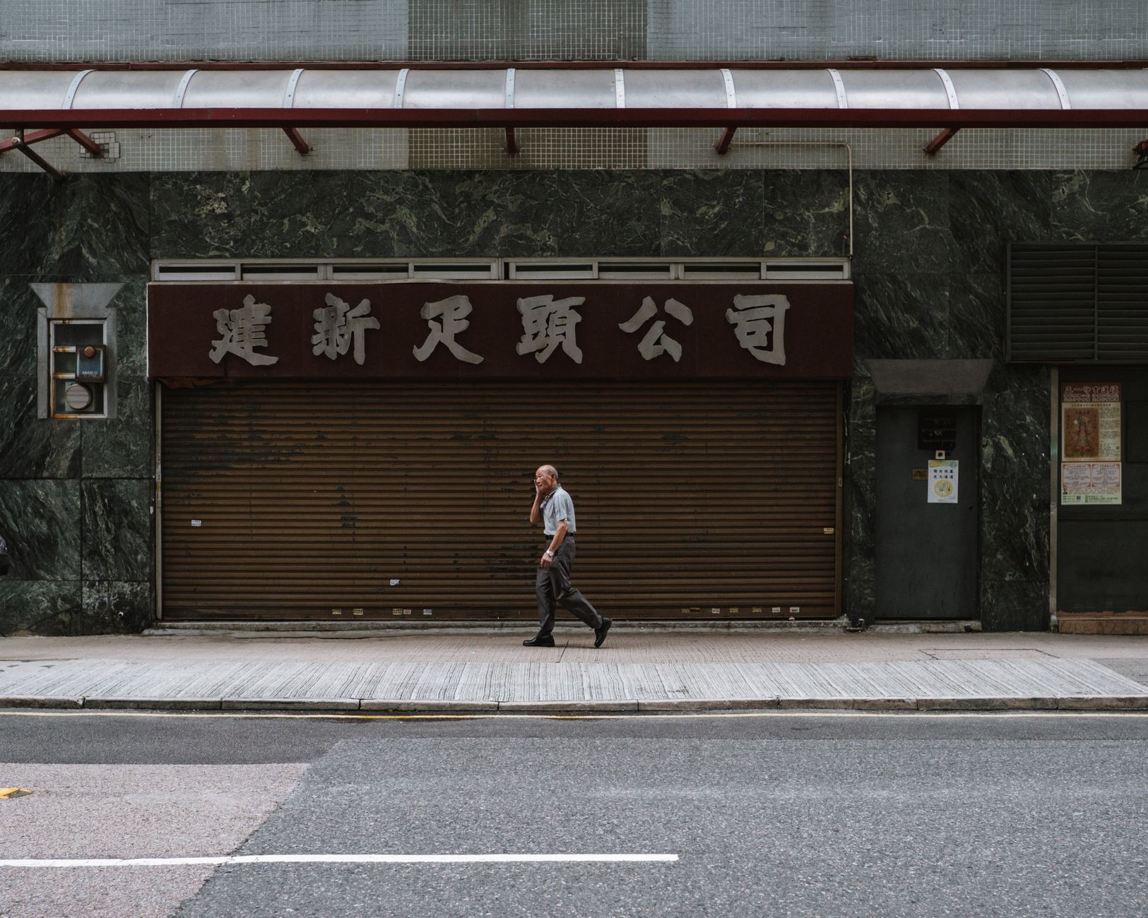 Elderly man walking past a closed shop with Chinese signage on a Hong Kong sidewalk.