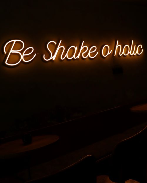 Free Illuminated neon sign displaying 'Be Shake o holic' on a dark background. Stock Photo