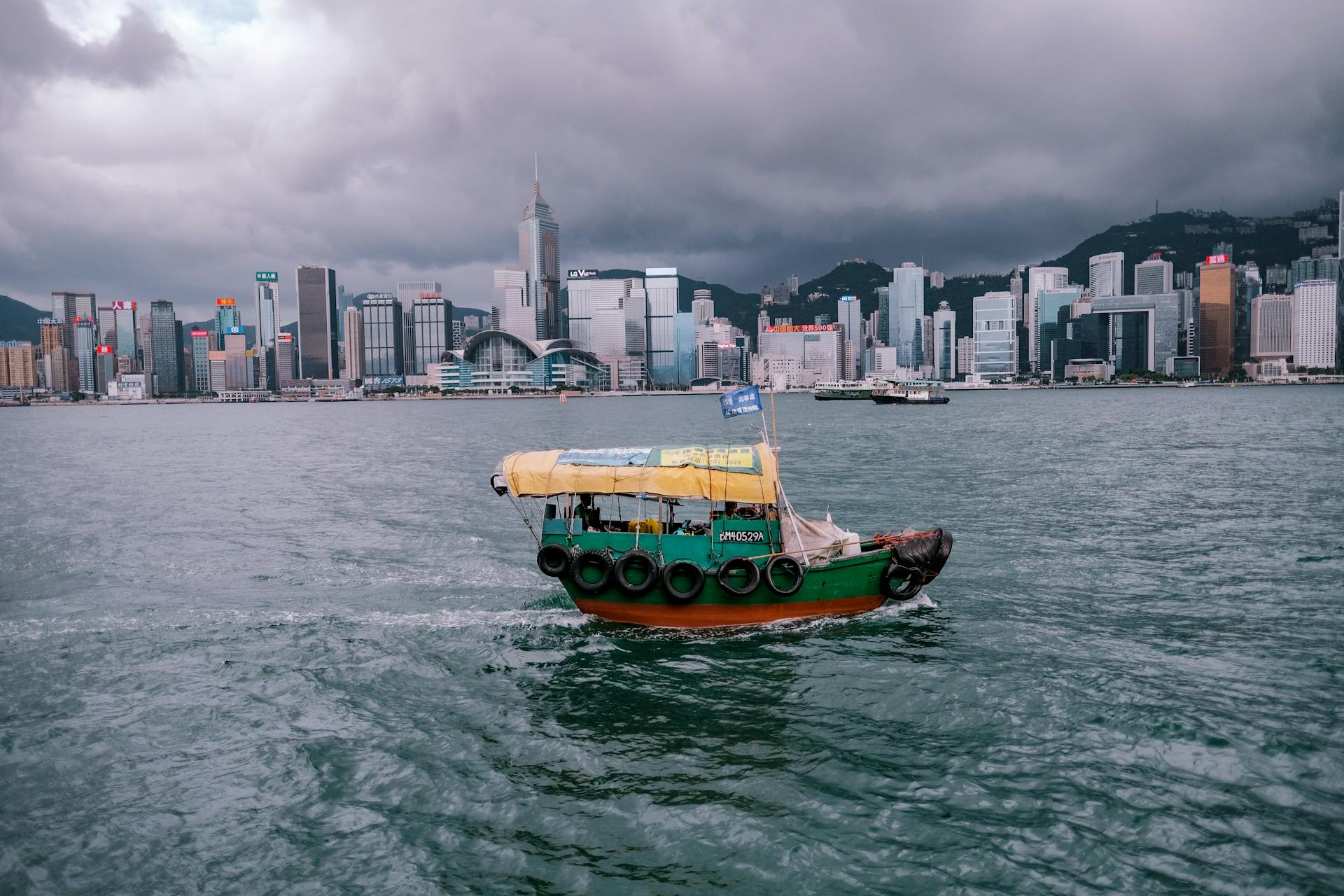 A traditional boat sails in Victoria Harbour with Hong Kong's iconic skyline under dramatic clouds.