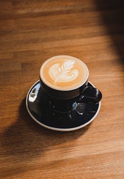 A beautifully crafted cappuccino with leaf latte art in a black ceramic cup on a wooden table.
