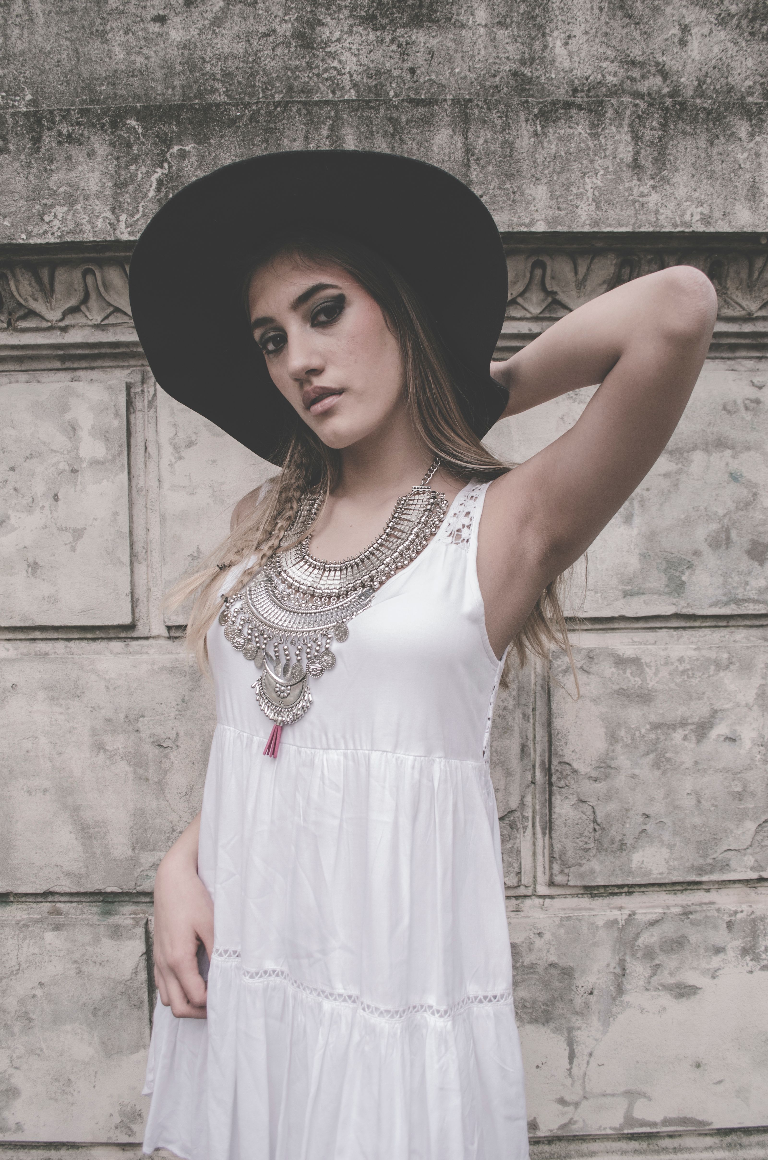 Mulher estilosa posando em um vestido branco e chapéu preto contra uma parede texturizada.