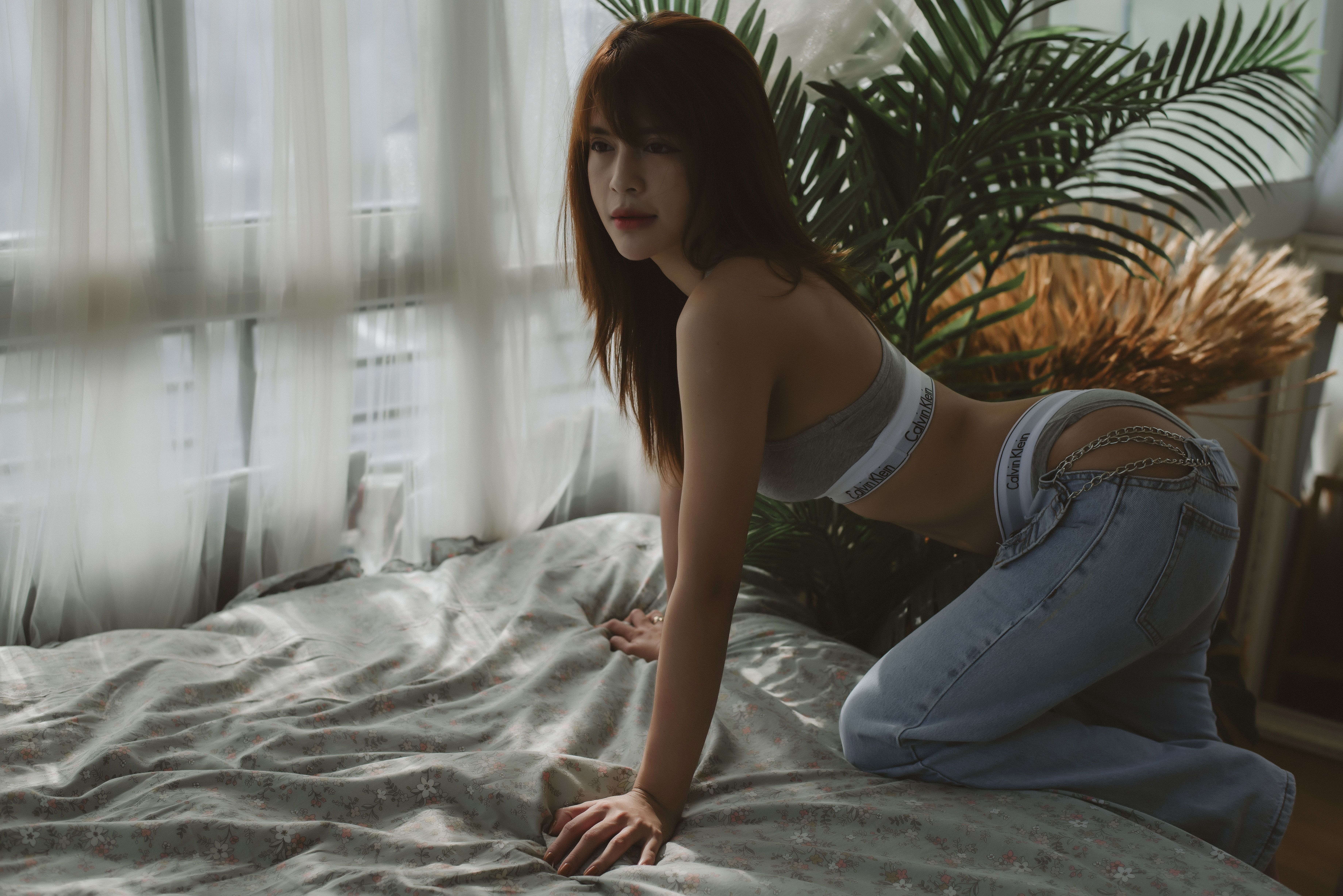 Woman in casual attire on bed, surrounded by natural light and foliage.