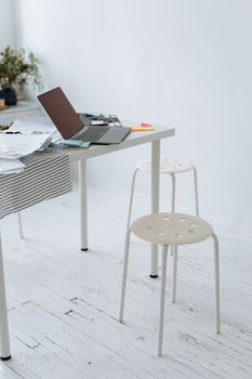 Minimalist home office setup with a laptop, papers, and stools on a white wooden floor.