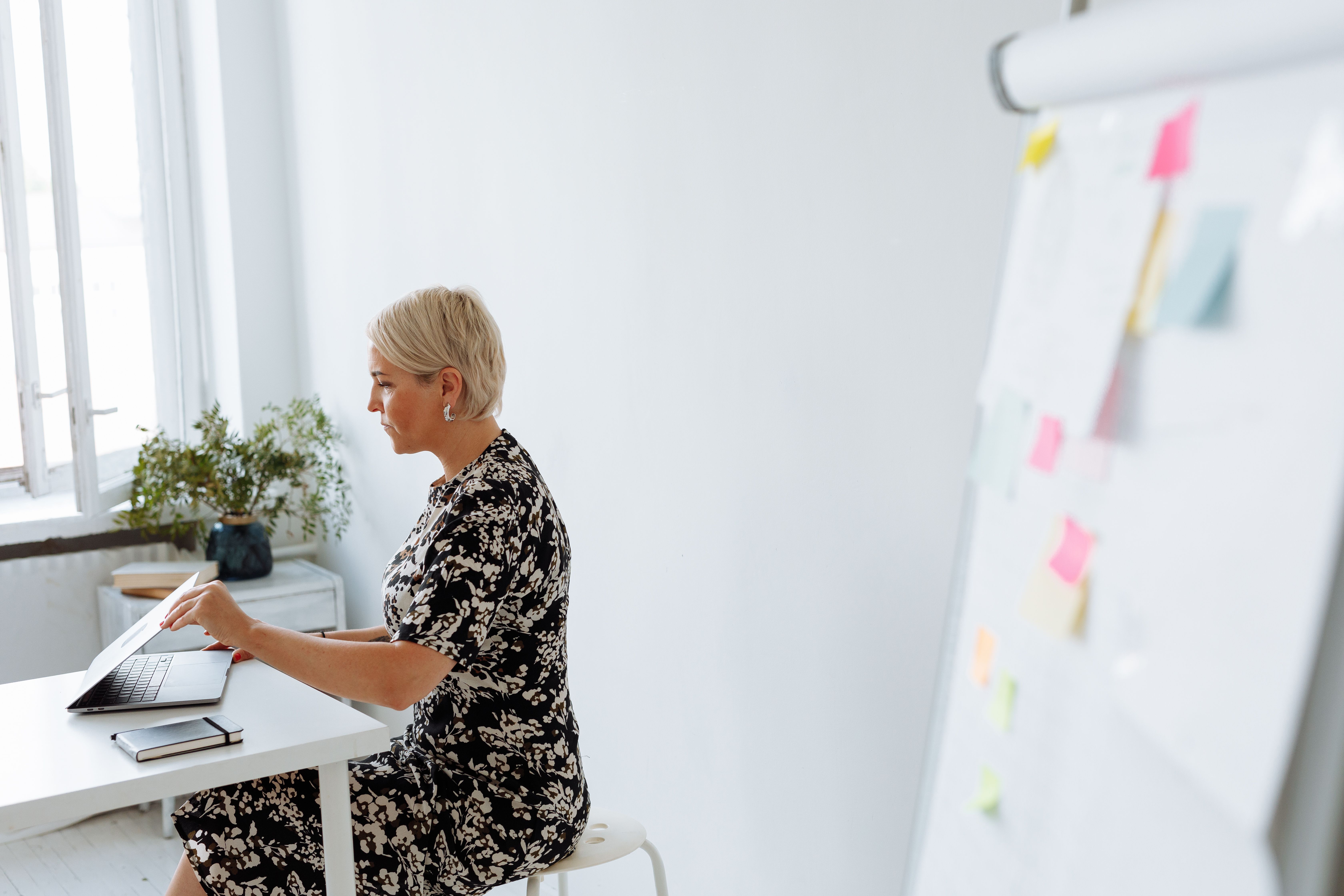 Middle-aged woman working on a laptop in a modern, minimalist office setting with a flip chart.