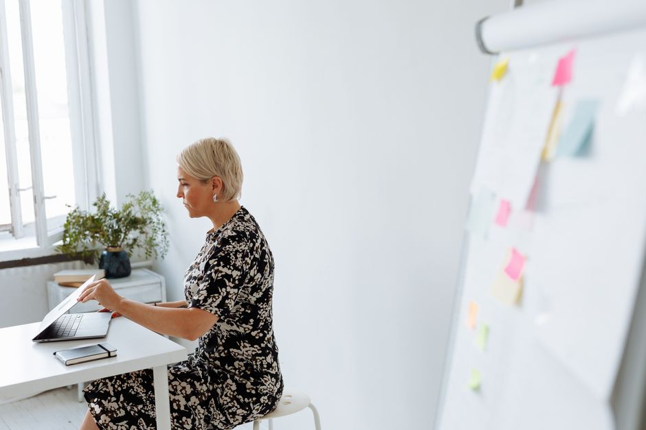 Middle-aged woman working on a laptop in a modern, minimalist office setting with a flip chart.