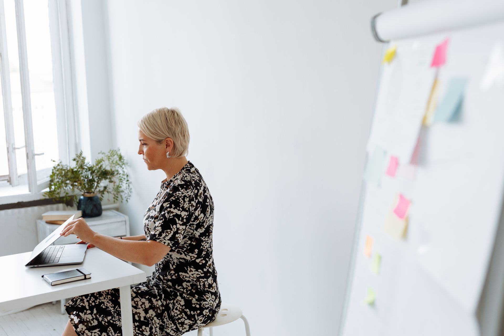 Middle-aged woman working on a laptop in a modern, minimalist office setting with a flip chart.