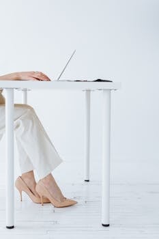 Chic workspace featuring a laptop on a white table with a person in stylish heels and white pants.