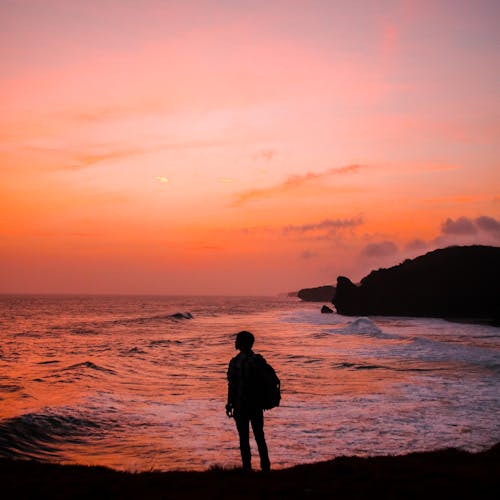 무료 황금 시간 동안 해변에 서있는 배낭을 가진 남자의 실루엣 사진 스톡 사진