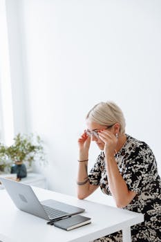 A woman adjusts her glasses while working on a laptop at a bright indoor workspace, embodying focus and productivity.