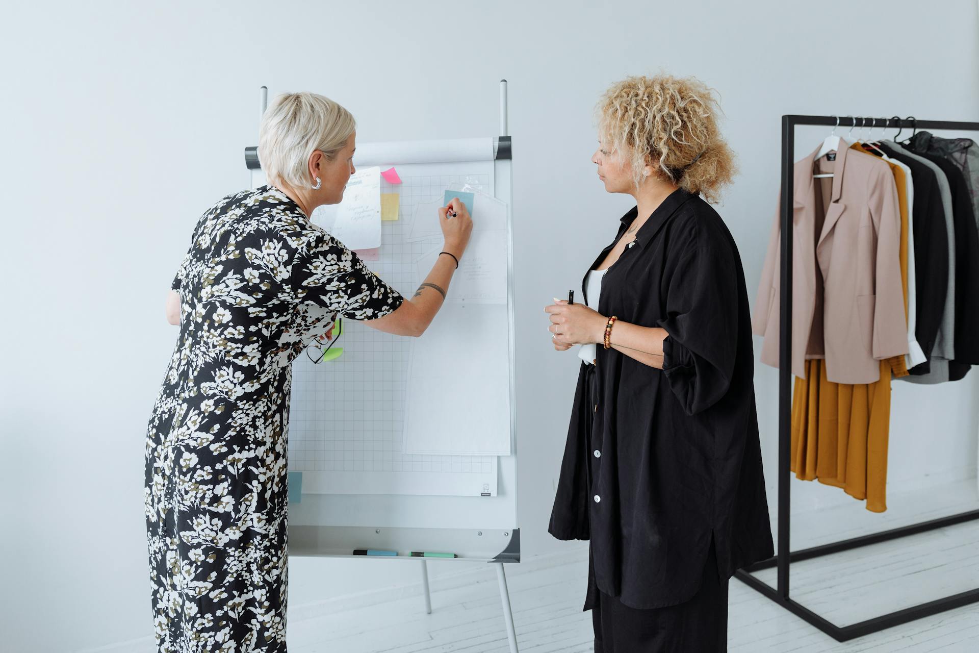 Two women work together on fashion concepts using a board and clothing rack.