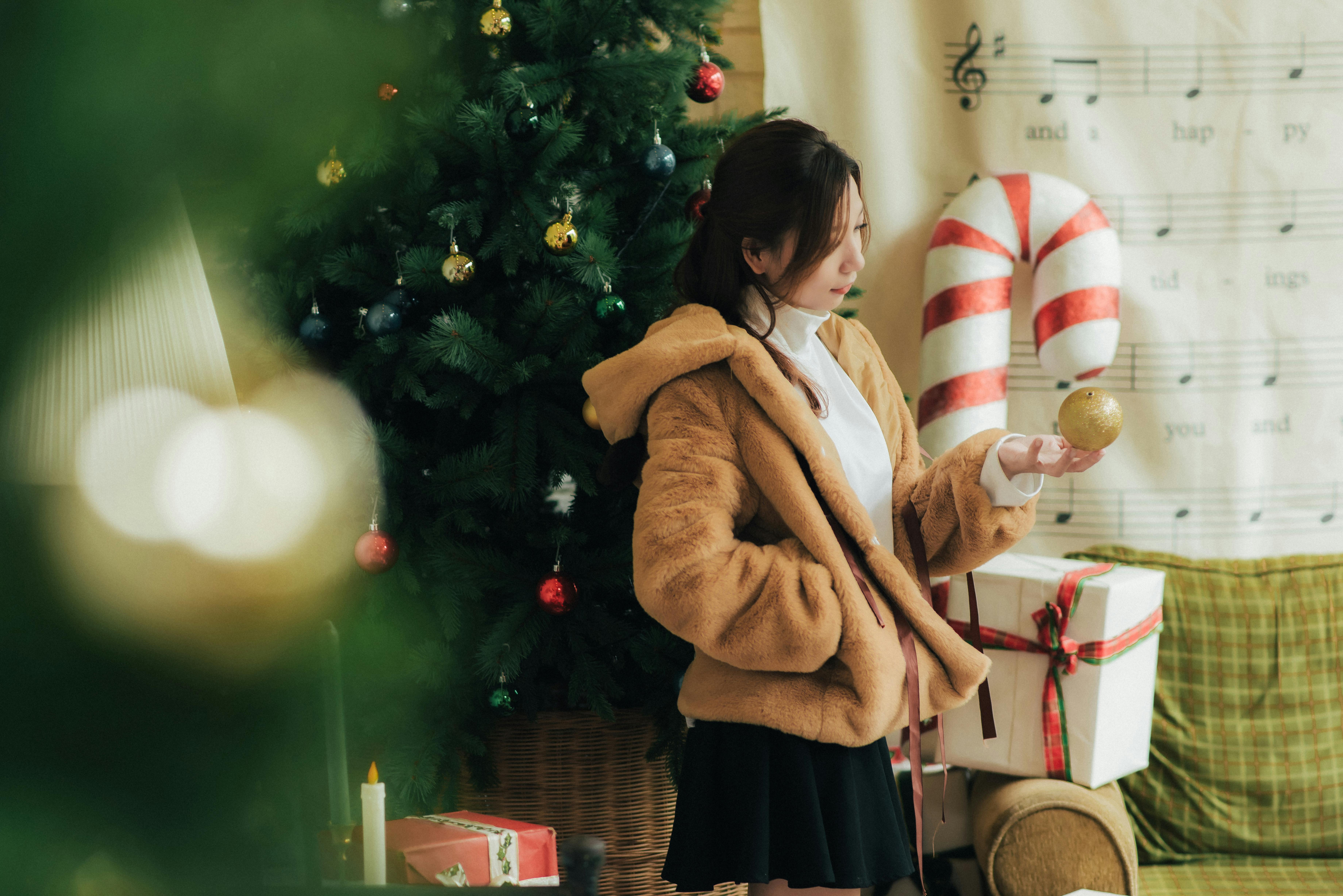 Young woman in fur coat by Christmas tree, holding an ornament indoors. Cozy and festive holiday scene.