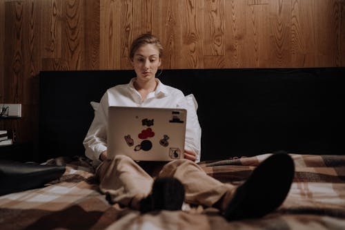Woman in White Long Sleeve Shirt Sitting on Bed while Using her Laptop