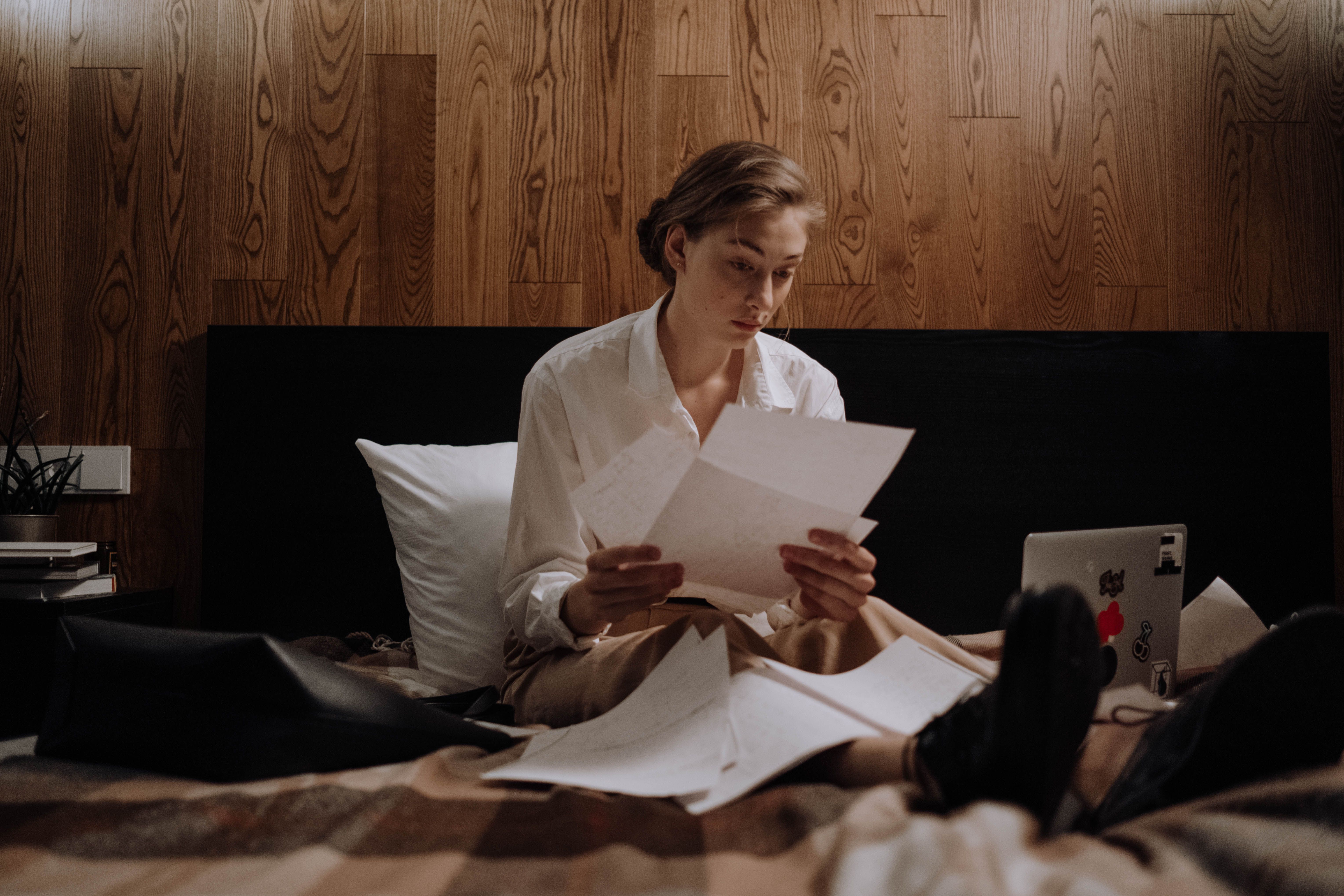 Woman sitting on bed, working from home with laptop and paperwork, focused and thoughtful.