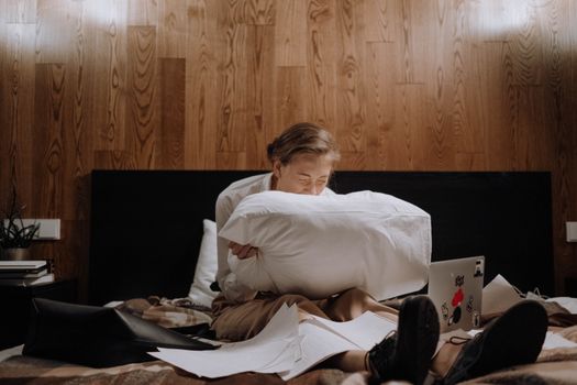 A woman sits alone on a bed, deep in thought, surrounded by papers and using a laptop.