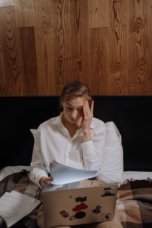 Free Woman feeling stressed working remotely, sitting in a cozy bedroom with papers and laptop. Stock Photo
